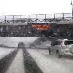 Cars driving in snow under winter storm warning sign