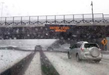 Cars driving in snow under winter storm warning sign