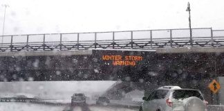 Cars driving in snow under winter storm warning sign