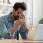 Man holding glasses, looking tired at laptop.