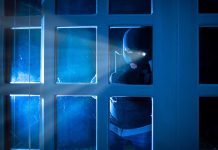 Masked figure peering through glass door at night.