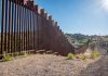 Tall metal border wall with rural landscape.