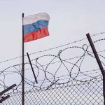 Barbed wire fence with Russian flag in background.