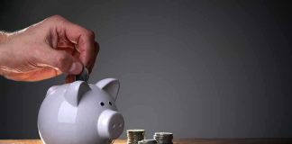 A hand inserting a coin into a white piggy bank on a wooden table with stacks of coins nearby