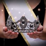 Sash-wearing person holding an ornate jeweled crown.