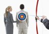 A woman places a target on a mans back while another man aims a bow and arrow at it
