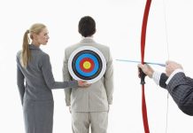 A woman places a target on a mans back while another man aims a bow and arrow at it