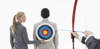 A woman places a target on a mans back while another man aims a bow and arrow at it