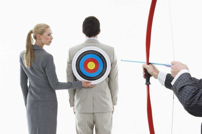 A woman places a target on a mans back while another man aims a bow and arrow at it