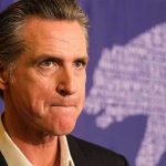 Resurfaced Clip IGNITES Newsom Culture War Man in suit, solemn expression, purple background with bear drawing.