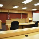 Firefighter’s Shocking Gun Attack—What Drove Him? Interior view of an empty courtroom with wooden furniture and American flags
