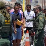 Border patrol officers speak with a line of people outdoors