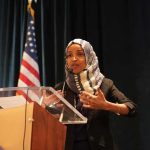Omar In Crosshairs For Fraud! Ilhan Omar speaking at a podium with a microphone