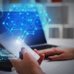 Zero‑Click Spyware Hits ICE — Encrypted Chats Exposed Hands using smartphone near laptop with digital hexagon interface overlay