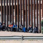41 Dead As Spain Overrun by Over 60K Illegals Group of people sitting by a tall border fence with an officer standing nearby