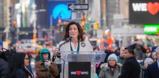 Kathy Hochul speaks at a clear podium in a city street; Josh Boyce stands among the crowd