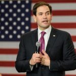 Iran Payday Looms — Rubio Races Gulf man in suit speaking with microphone before American flag