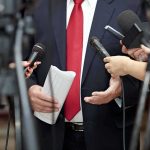 Republican Revolt Hits Trump’s Intel Pick Man in suit with red tie speaking to reporters holding microphones