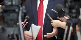 Man in suit with red tie speaking to reporters holding microphones