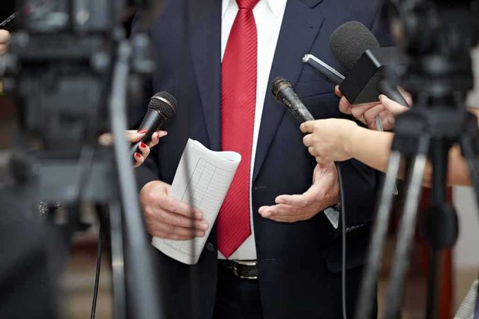 shutterstock_82082848 Man in suit with red tie speaking to reporters holding microphones
