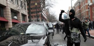 Masked protester raises tool to strike black car on city street